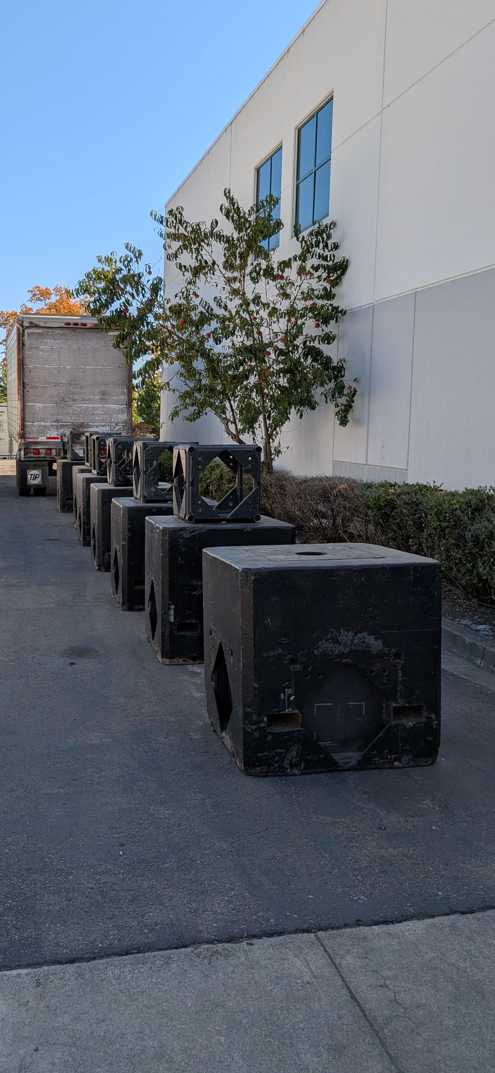 Row of large, black shipping boxes lined up on a paved area beside a building, with a delivery truck parked in the background.