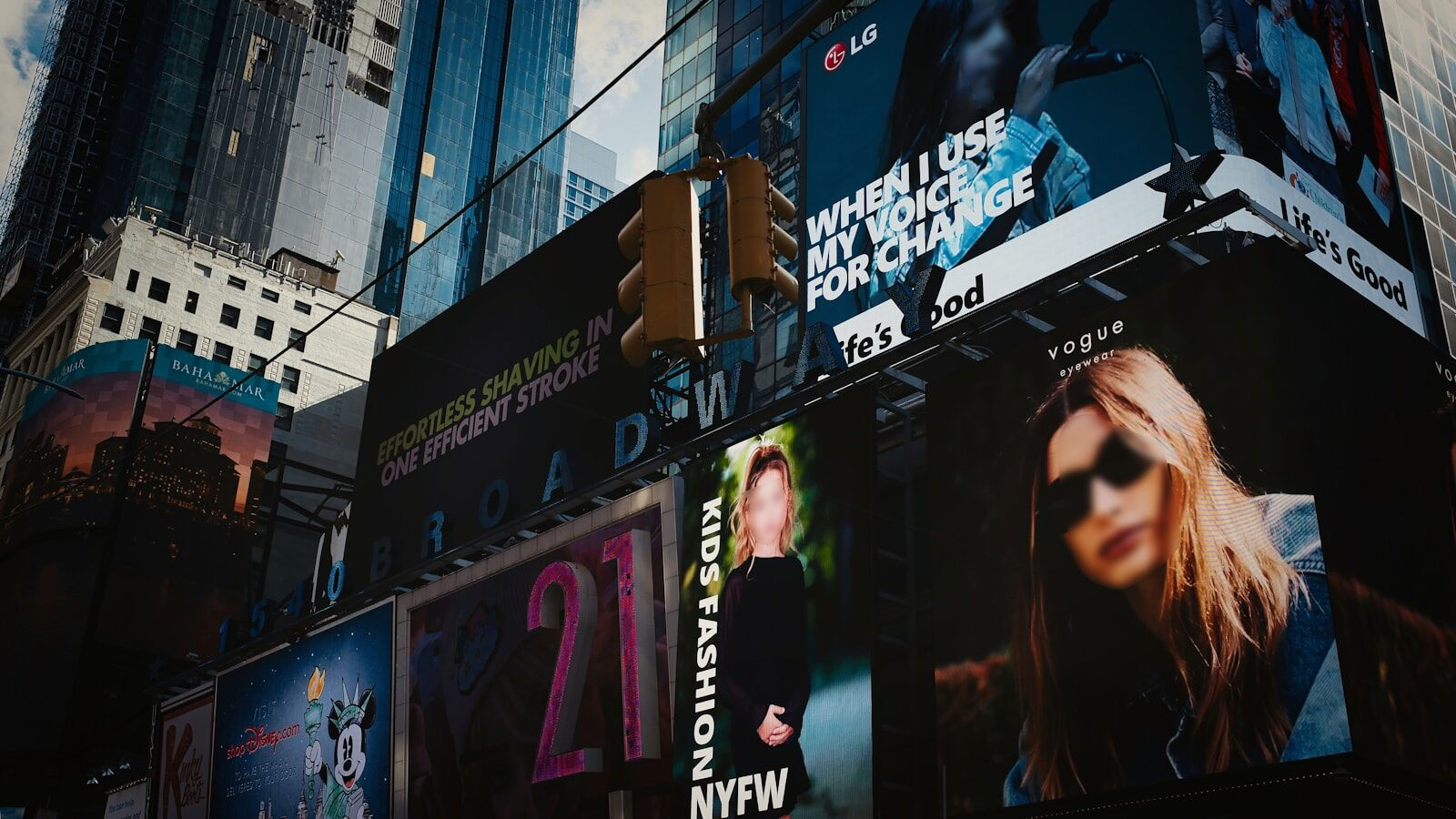 Brightly lit Times Square showcasing various advertisements, including fashion promotions for NYFW and eyewear by Vogue, under a blue sky.